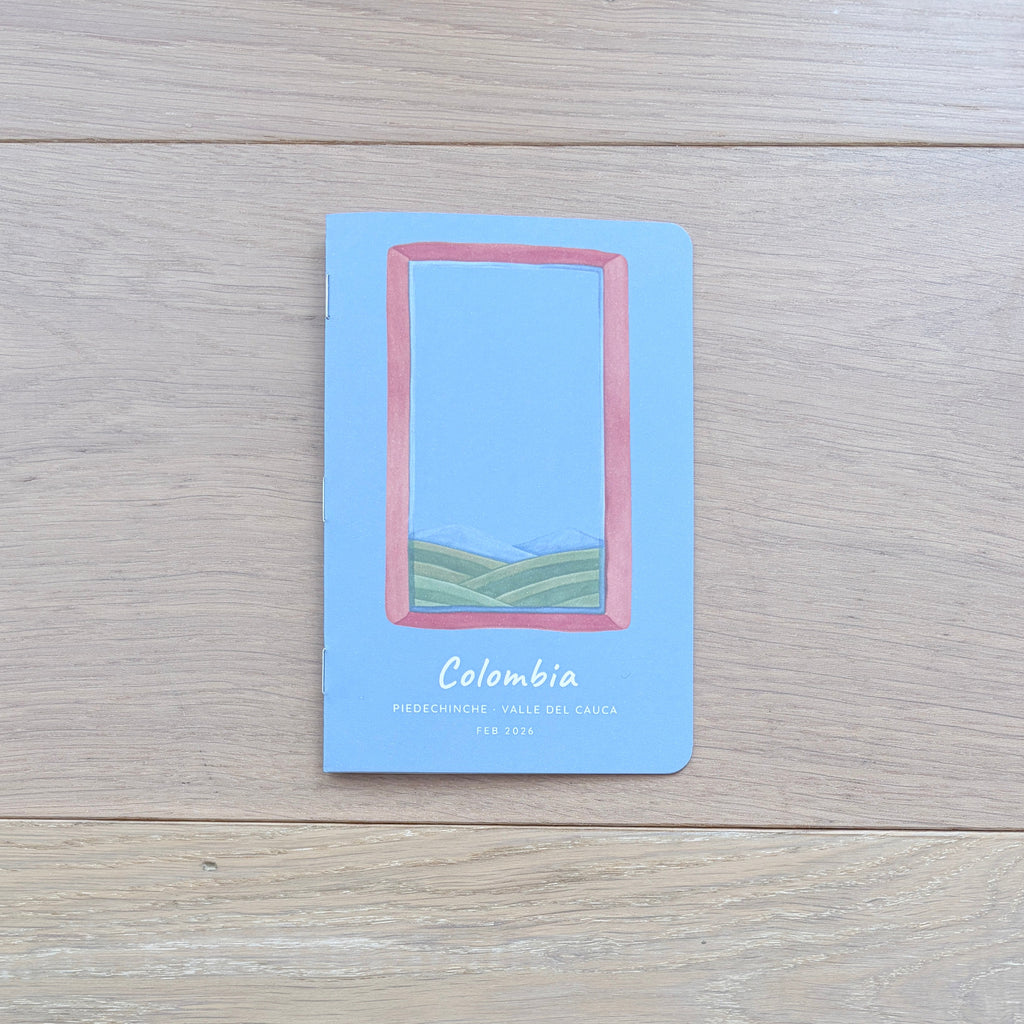 A small travel notebook or card featuring a landscape illustration labeled 'Colombia' sits on a light wood tabletop. The cover is light blue with a pink rectangular border and a small green landscape scene.
