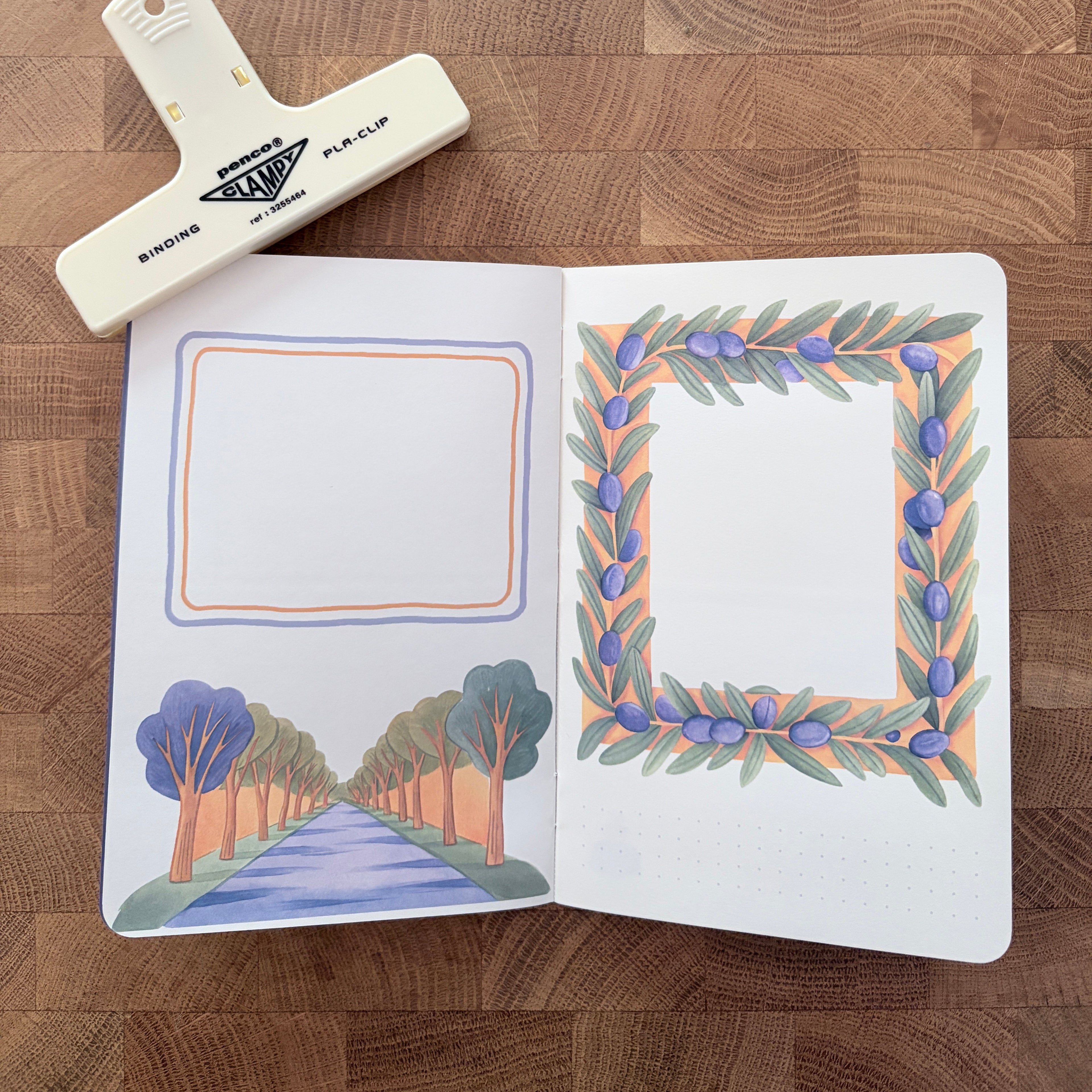 An open bullet journal or notebook on a wooden tabletop, featuring decorative watercolor frame illustrations on the pages. A white plastic binder clip labeled 'BINDER PLACHIP' rests above the notebook.
