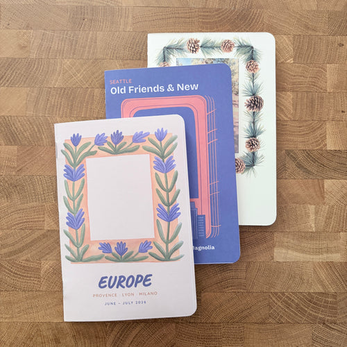 Three decorative paper notepads or cardstock pads arranged on a wood-grain tabletop. The front pad is white with a floral border and the word EUROPE; the middle pad is blue with a pink frame design and partial text reading Old Friends & New; the back pad is white with a pine and berry border.