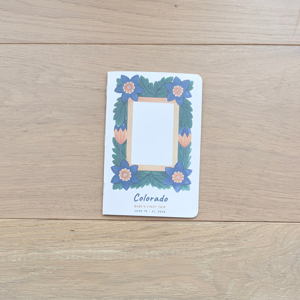 A small printed card or notebook cover with a floral border and the word 'Colorado' sits on a light wood tabletop. The product is photographed in a real environment, with part of a person's legs and a tripod visible underneath the table.