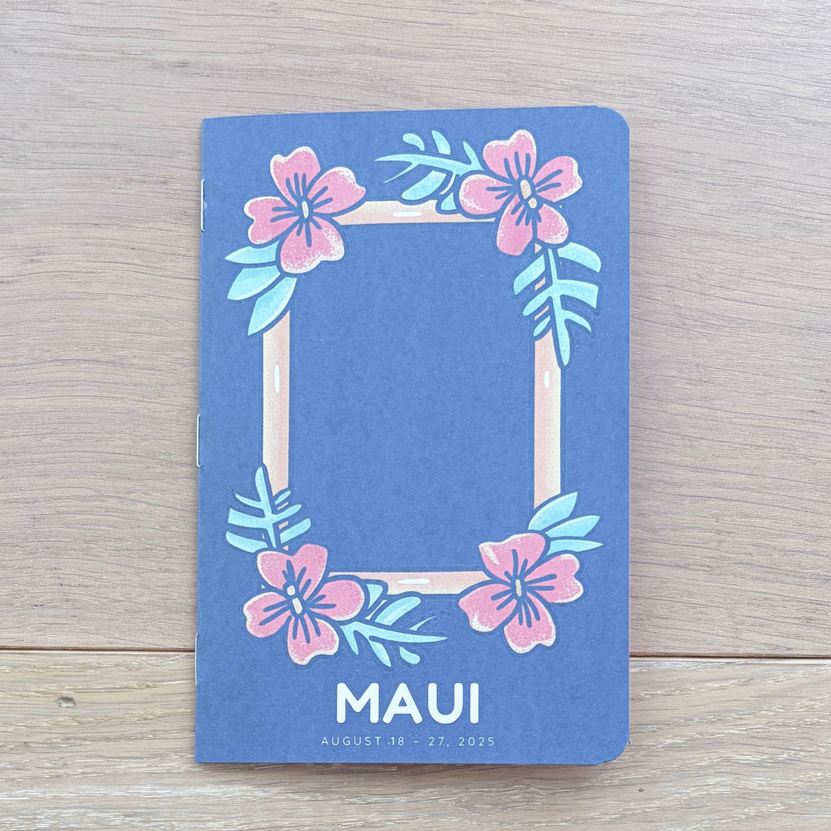 A blue rectangular card or notebook cover with a floral frame design and the word 'MAUI' at the bottom, placed on a light wood tabletop. Parts of a person's legs and clothing are visible underneath the table.