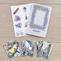 A set of Europe-themed travel cards and matching photo prints arranged on a light wood tabletop. The cards feature illustrated motifs like a bicycle, coffee cup, and bread, with one card displaying a colorful leaf wreath border and the word 'EUROPE'.