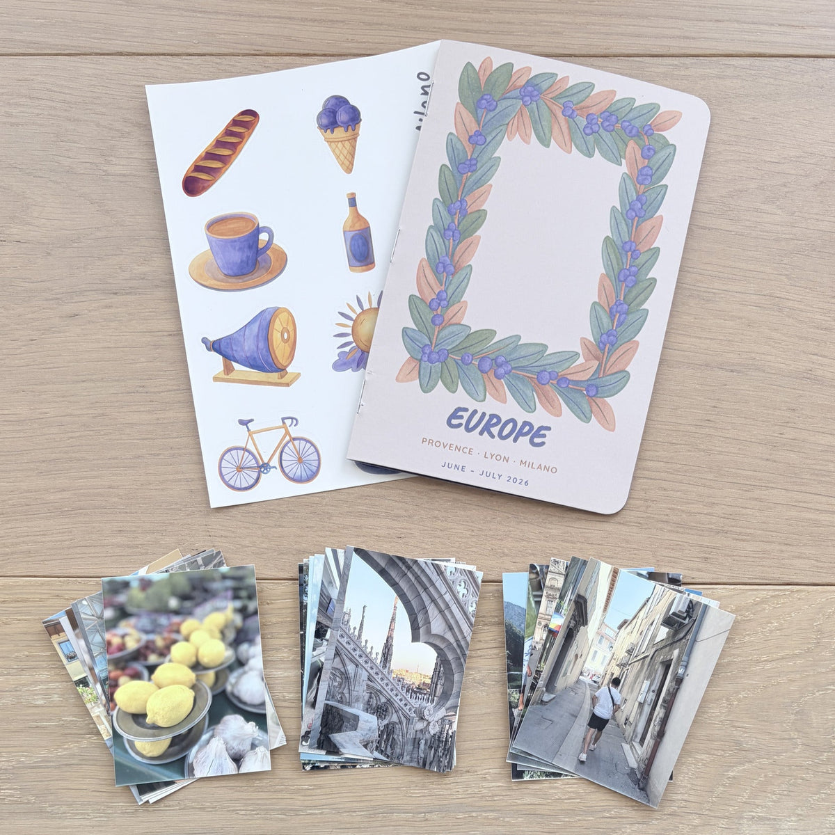 A set of Europe-themed travel cards and matching photo prints arranged on a light wood tabletop. The cards feature illustrated motifs like a bicycle, coffee cup, and bread, with one card displaying a colorful leaf wreath border and the word 'EUROPE'.