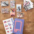 A styled flat-lay of printed stationery and photo prints arranged on a wooden tabletop. The set includes a purple notebook with a pink graphic, a white sheet with colorful Seattle-themed illustrations and the word 'Seattle,' several small printed photos, and a stack of photo prints.