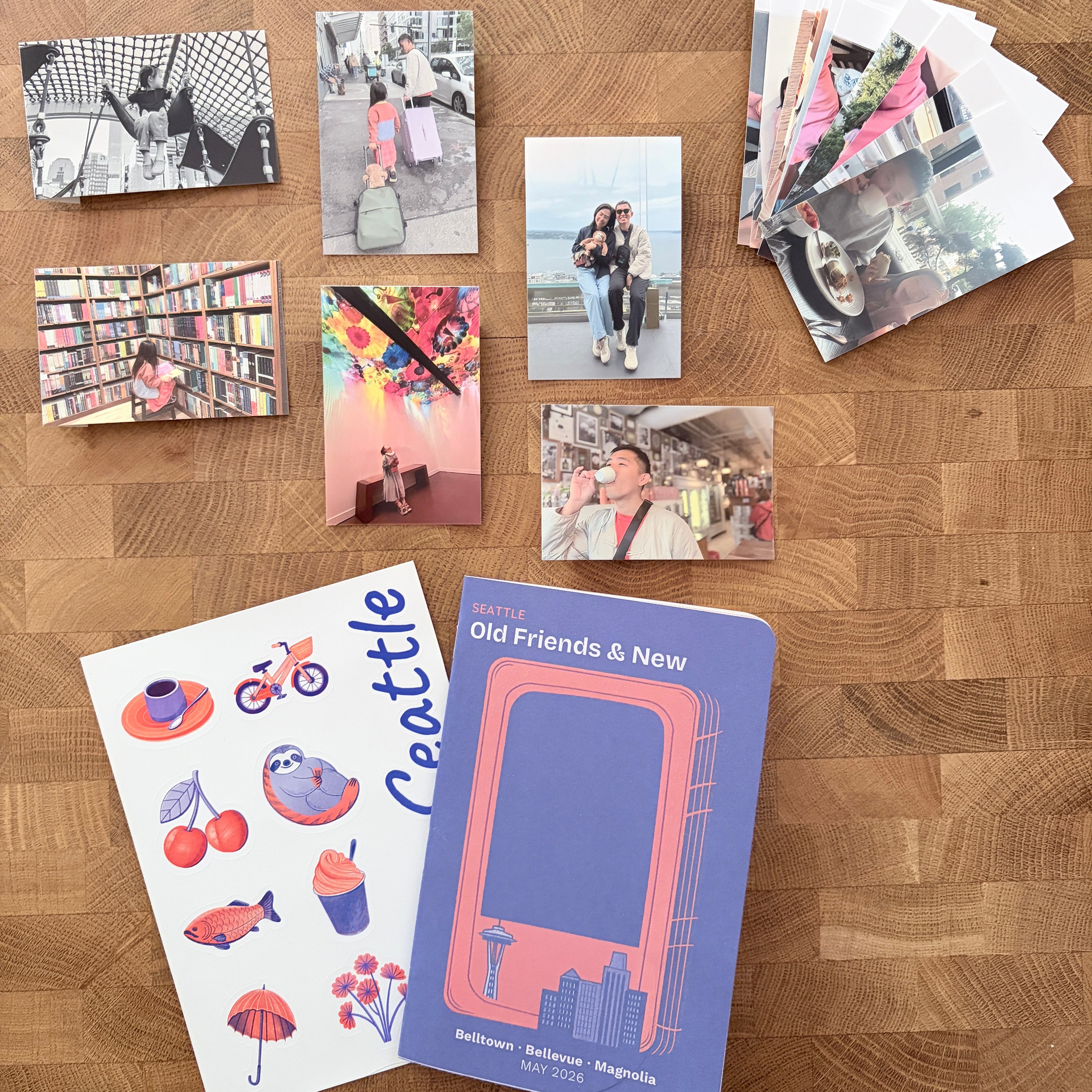 A styled flat-lay of printed stationery and photo prints arranged on a wooden tabletop. The set includes a purple notebook with a pink graphic, a white sheet with colorful Seattle-themed illustrations and the word 'Seattle,' several small printed photos, and a stack of photo prints.