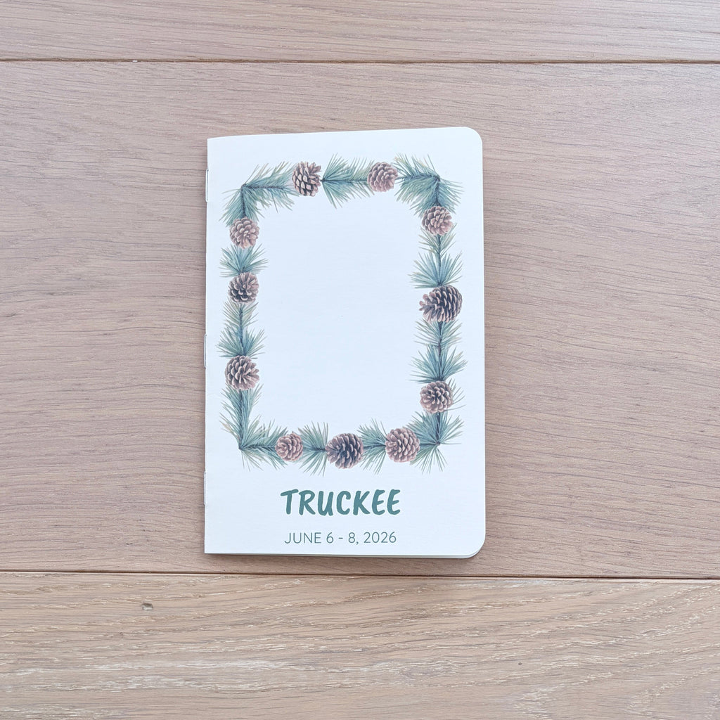 A white hardcover notebook or journal titled 'TRUCKEE' with a pinecone and evergreen wreath border, placed on a light wood tabletop. The date 'JUNE 9, 2021' is printed below the title.